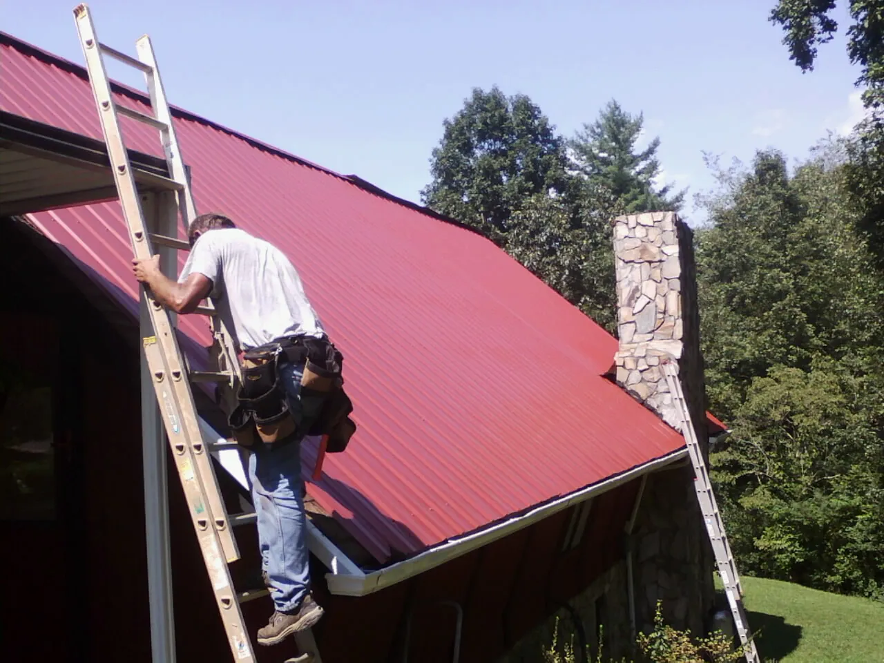 Roofer on a ladder beside a red metal roof and stone chimney — Soffit & Fascia Repair by Roofing Brothers in Dale
